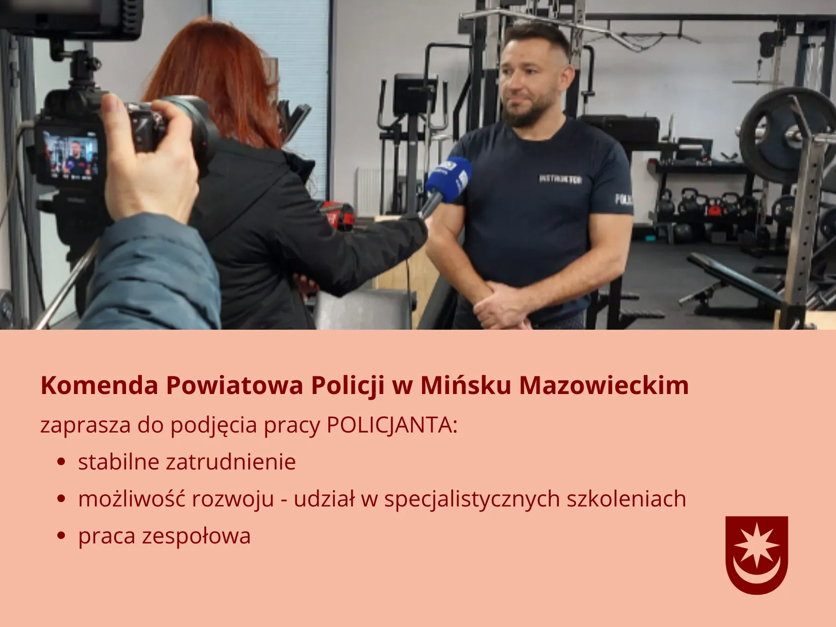 Mężczyzna w ciemnej koszulce stoi na siłowni i udziela wywiadu. Kobieta z czerwonymi włosami trzyma mikrofon, a z lewej strony widać kamerę. W tle widoczne są maszyny i hantle na siłowni. Na dole obrazka znajduje się różowy baner z informacjami rekrutacyjnymi i herbem.