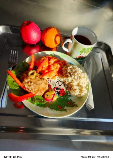 Metalowa taca z talerzem w dziecięcy wzór. Na talerzu leży panierowany kotlet, porcja kuskusu, ziemniaki z sosem marchewkowym, liść sałaty i paski czerwonej papryki. Na tacy obok stoi biały kubek z ciemnym napojem. Obok leżą czerwone jabłko i pomarańcza. Widelec i nóż leżą po bokach tacy. Jasne naturalne światło oświetla scenę.