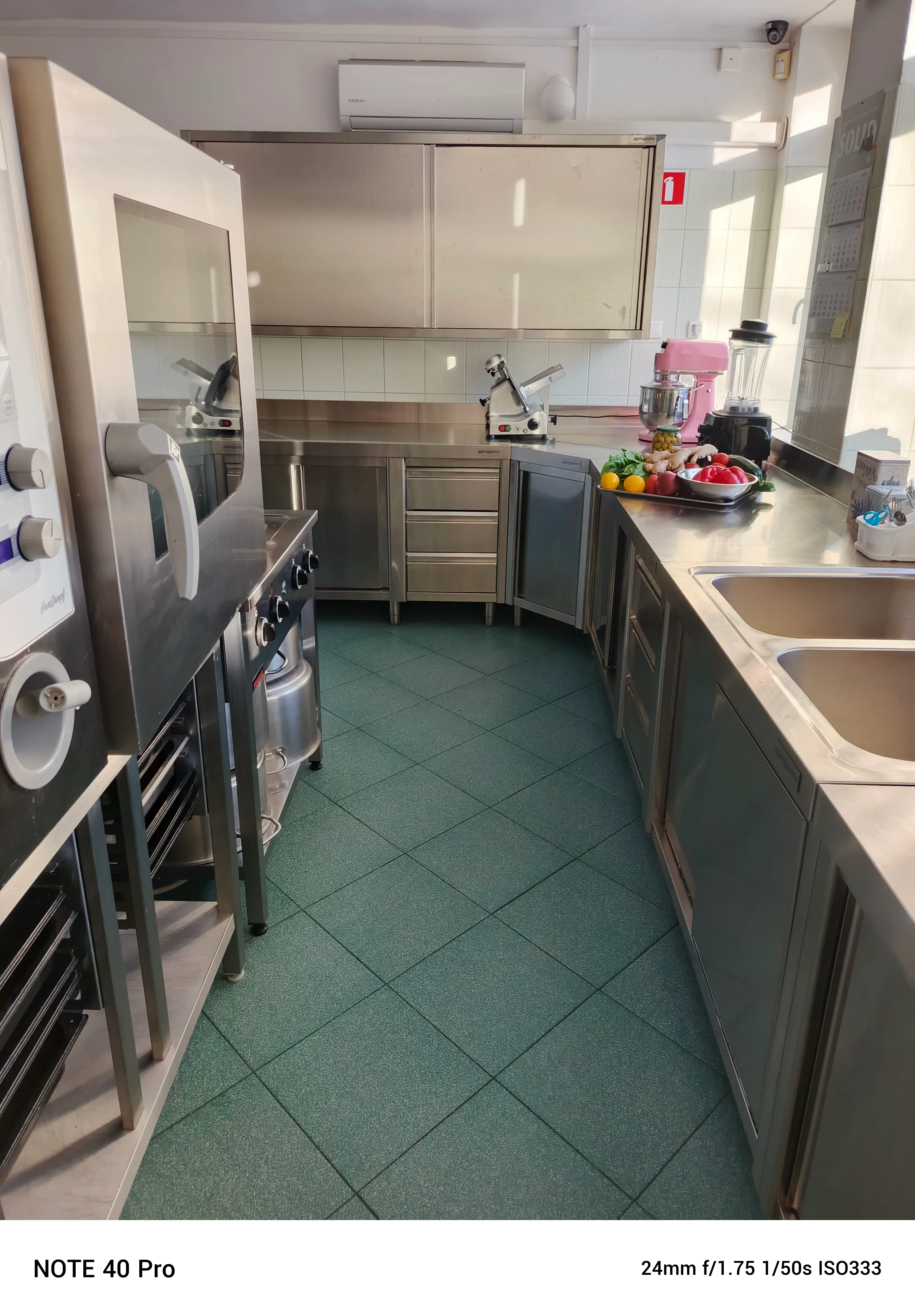 Profesjonalna kuchnia ze stalowymi blatami i szafkami. Zielona kafelkowa podłoga i piec po lewej stronie. Na prawym blacie stoi robot kuchenny, blender i taca z warzywami. Okno wpuszcza dzienne światło.