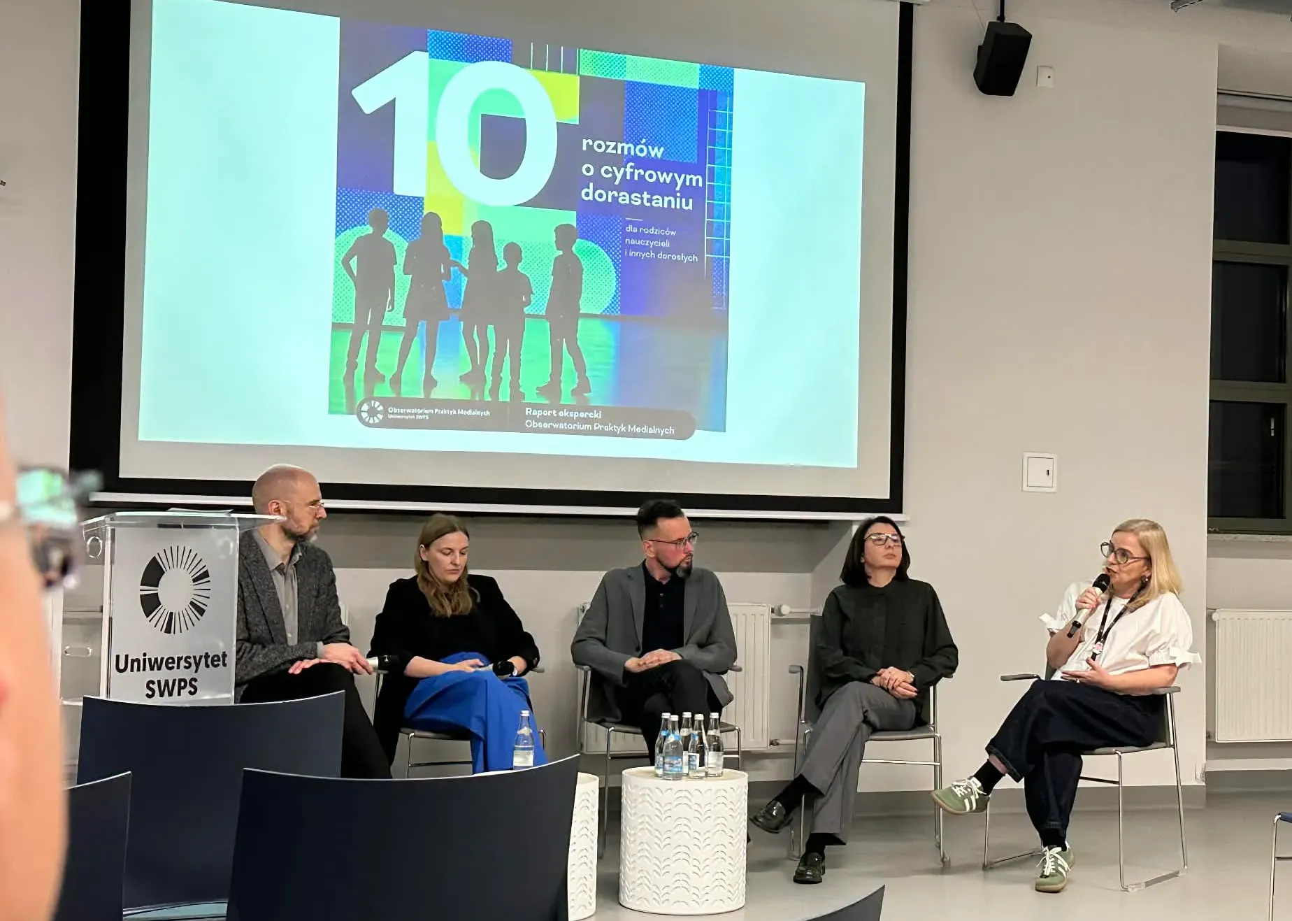 Sala wykładowa z panelem pięciu osób siedzących w rzędzie przed ekranem projekcyjnym. Ekran wyświetla kolorową grafikę informacyjną. Po lewej stoi przezroczyste podium z logo uczelni. Paneliści siedzą na krzesłach; prawa osoba trzyma mikrofon i mówi. W pomieszczeniu jasne ściany, okno z prawej i kilka pustych krzeseł publiczności z przodu.