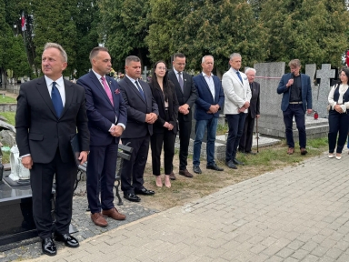 Dziesięć osób stoi na zewnątrz na cmentarzu przy pomnikach i nagrobkach, jedna osoba mówi do mikrofonu.