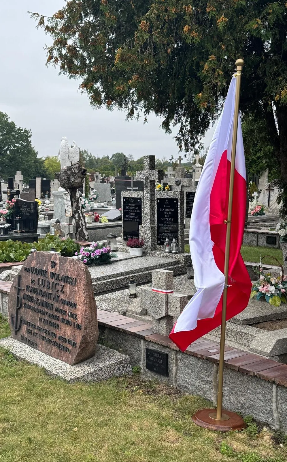 Pochmurny dzień na cmentarzu z pomnikiem i stojącą obok flagą Polski na tle wielu grobów i zielonej trawy.