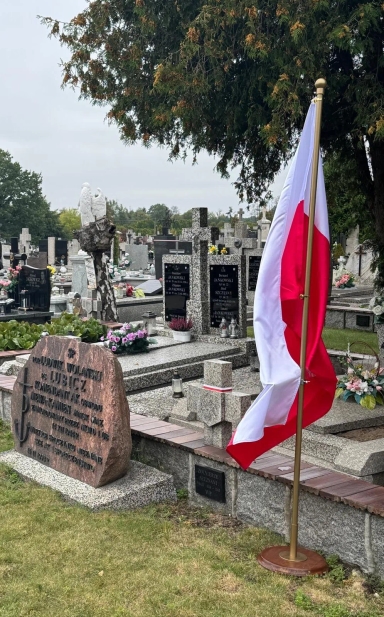Pochmurny dzień na cmentarzu z pomnikiem i stojącą obok flagą Polski na tle wielu grobów i zielonej trawy.