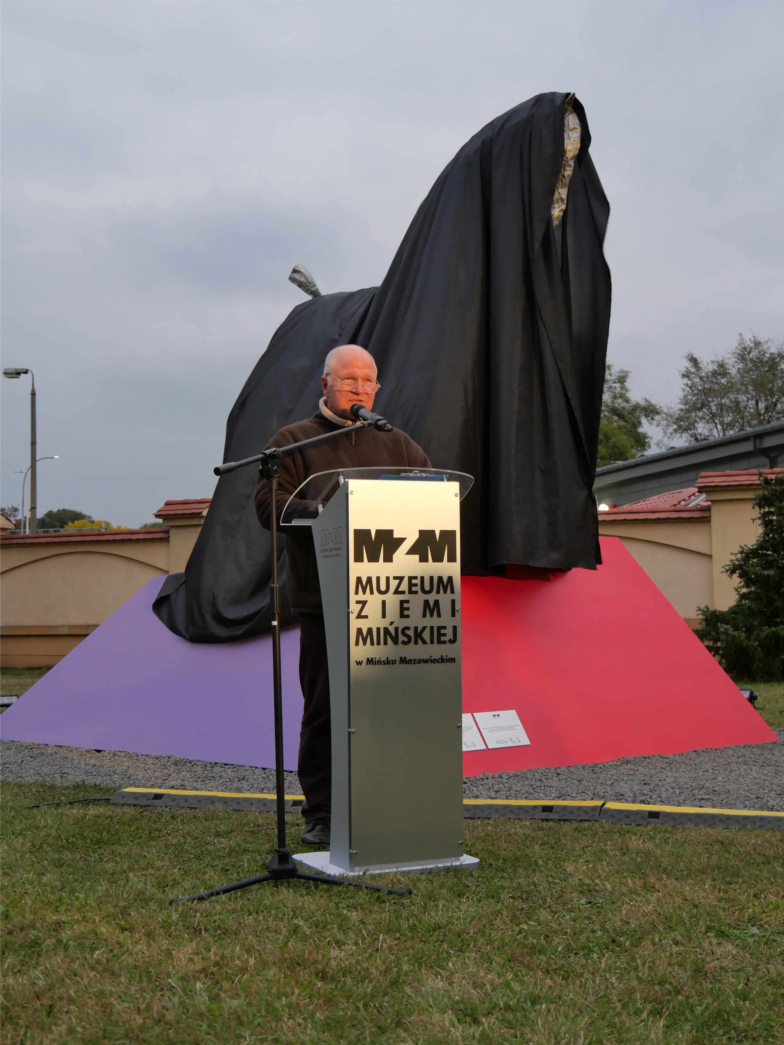 Mężczyzna przemawia przy mównicy na zewnątrz, za nim zasłonięta czarnym płótnem duża rzeźba na podstawie w kolorach fioletowym i czerwonym, trawa i budynki w tle.