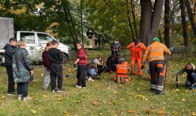 Grupa ludzi na trawie w parku jesienią. Kilka osób klęczy i pracuje przy ziemi, dwie osoby ubrane w pomarańczowe ubrania robocze, inni stoją lub kucają. W tle białe auto dostawcze oraz drzewa z zielonymi i żółtymi liśćmi.