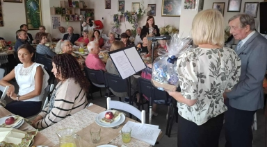 Kilka osób siedzi przy stołach z jedzeniem wewnątrz pomieszczenia. Dwie osoby stoją przy mikrofonie. Jedna trzyma prezent.