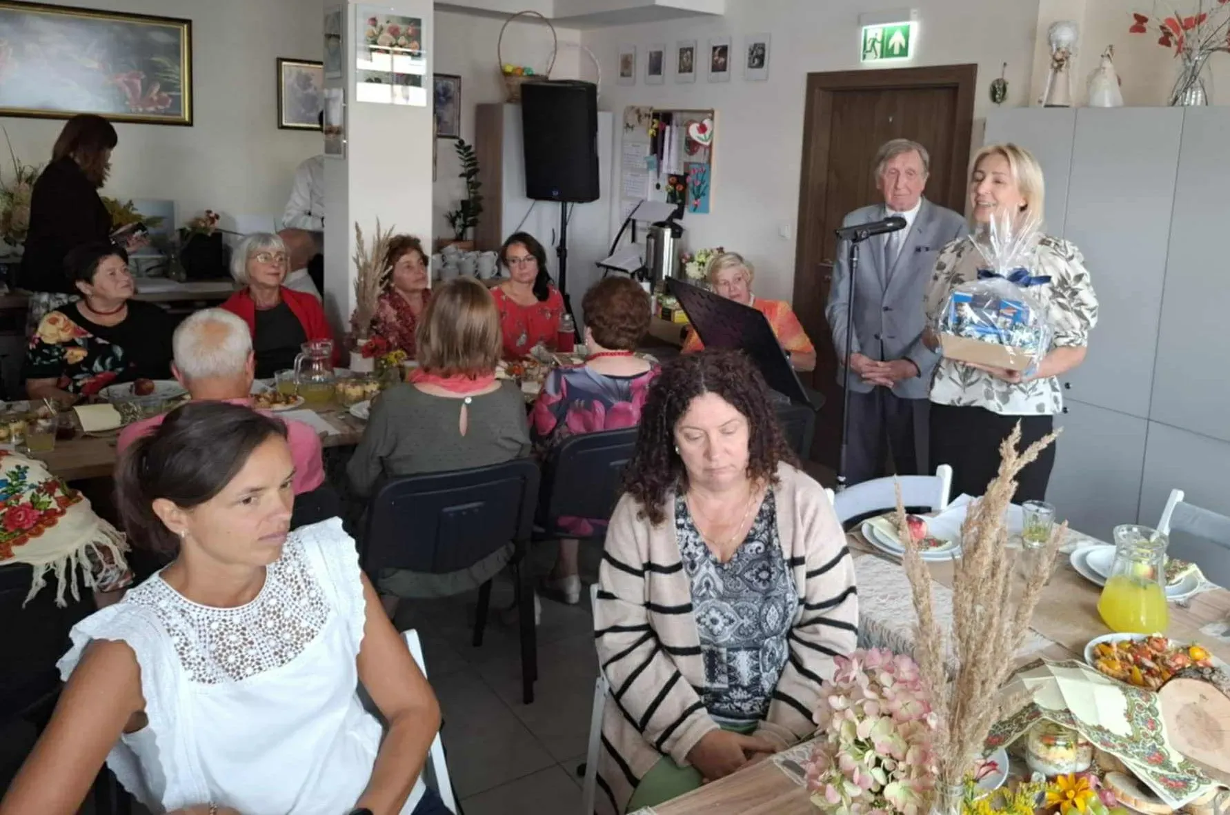 Grupa dorosłych osób siedzi przy stołach w pomieszczeniu. Kobieta przy mikrofonie trzyma podarunek, mężczyzna stoi obok niej.
