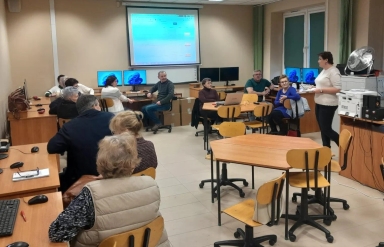 Grupa dorosłych osób siedzi w sali komputerowej na krzesłach przy stolikach. Kobieta stoi obok biurka po prawej stronie i zwraca się do pozostałych. W tle projektor wyświetla obraz na ekranie, a przy ścianie stoją komputery. Pokój jest jasno oświetlony, na podłodze jasne płytki.