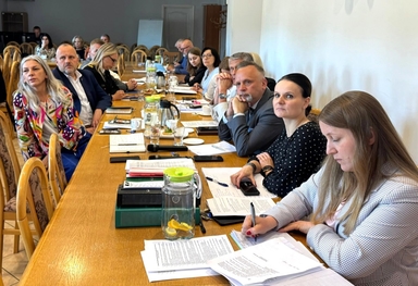 Około tuzina osób siedzi przy długim drewnianym stole wewnątrz sali konferencyjnej. Na stole leżą dokumenty, notatniki, filiżanki i dzbanki z wodą i plasterkami cytryny, a uczestnicy słuchają i notują.