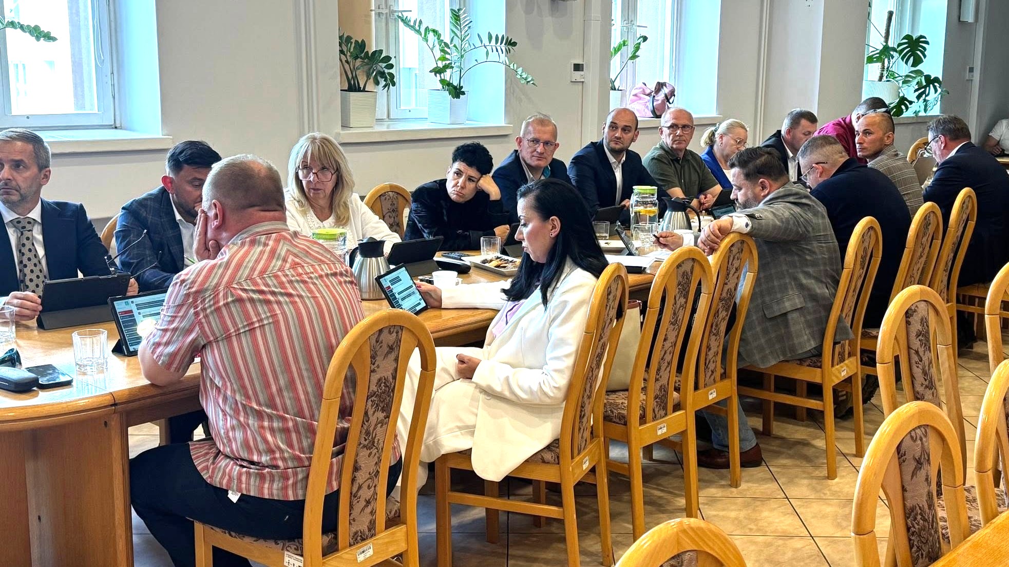 Około piętnastu dorosłych siedzi przy długim drewnianym stole w jasnej sali konferencyjnej. Okna z doniczkami roślin wpuszczają dzienne światło. Na stole stoją laptopy, tablety, termosy i szklanki. Kobieta w białym garniturze siedzi na pierwszym planie, inni rozmawiają lub patrzą na urządzenia.