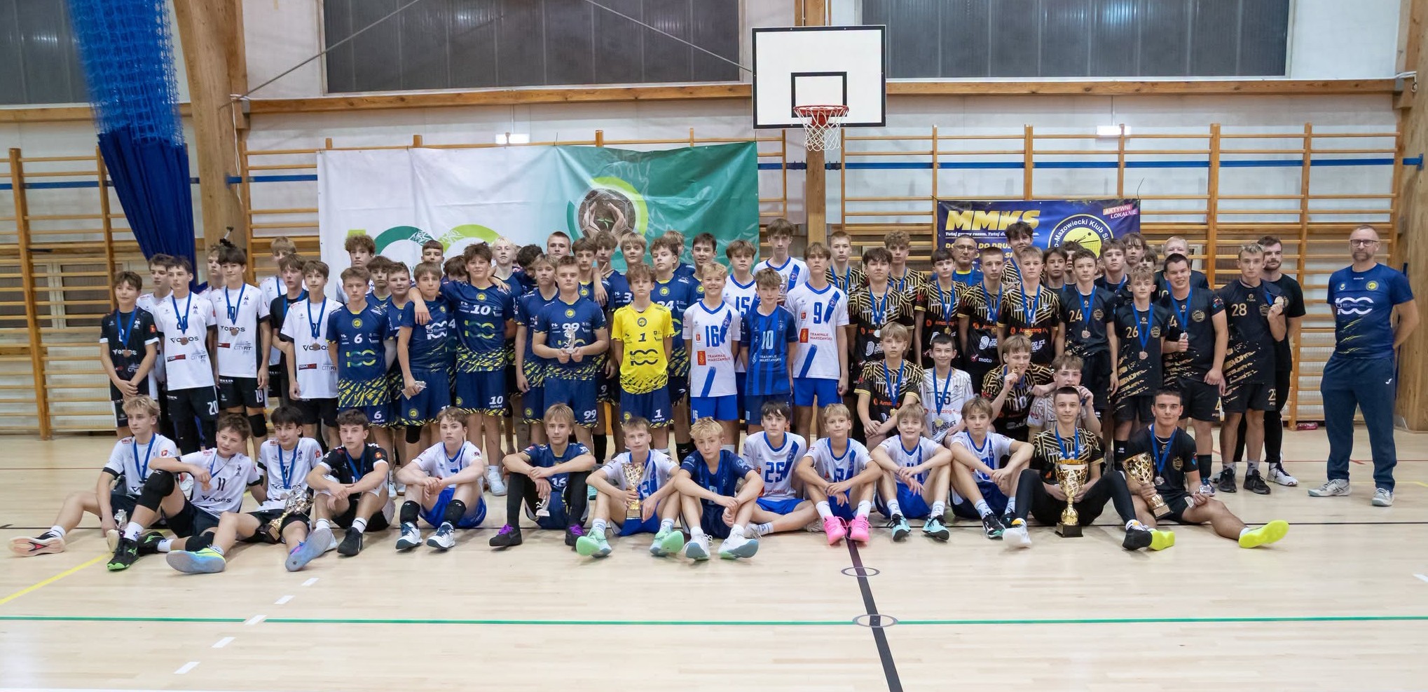 Duża grupa młodych zawodników siatkówki i trenerów stoi i siedzi w hali sportowej pod koszem. Drużyny mają różnokolorowe stroje (białe, granatowe, niebieskie, czarne) i trzymają medale oraz puchary na drewnianej podłodze przed ścianą z drabinkami.