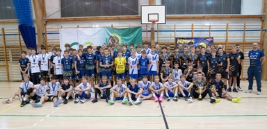 Duża grupa młodych zawodników siatkówki i trenerów stoi i siedzi w hali sportowej pod koszem. Drużyny mają różnokolorowe stroje (białe, granatowe, niebieskie, czarne) i trzymają medale oraz puchary na drewnianej podłodze przed ścianą z drabinkami.
