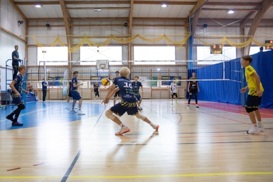 Hala sportowa z boiskiem do siatkówki i drewnianą konstrukcją sufitu. Grupa młodych zawodników gra przy siatce, piłka unosi się nad środkiem boiska. Sędzia stoi na podwyższeniu po lewej stronie. Niebieskie zasłony i tablica wyników w tle.