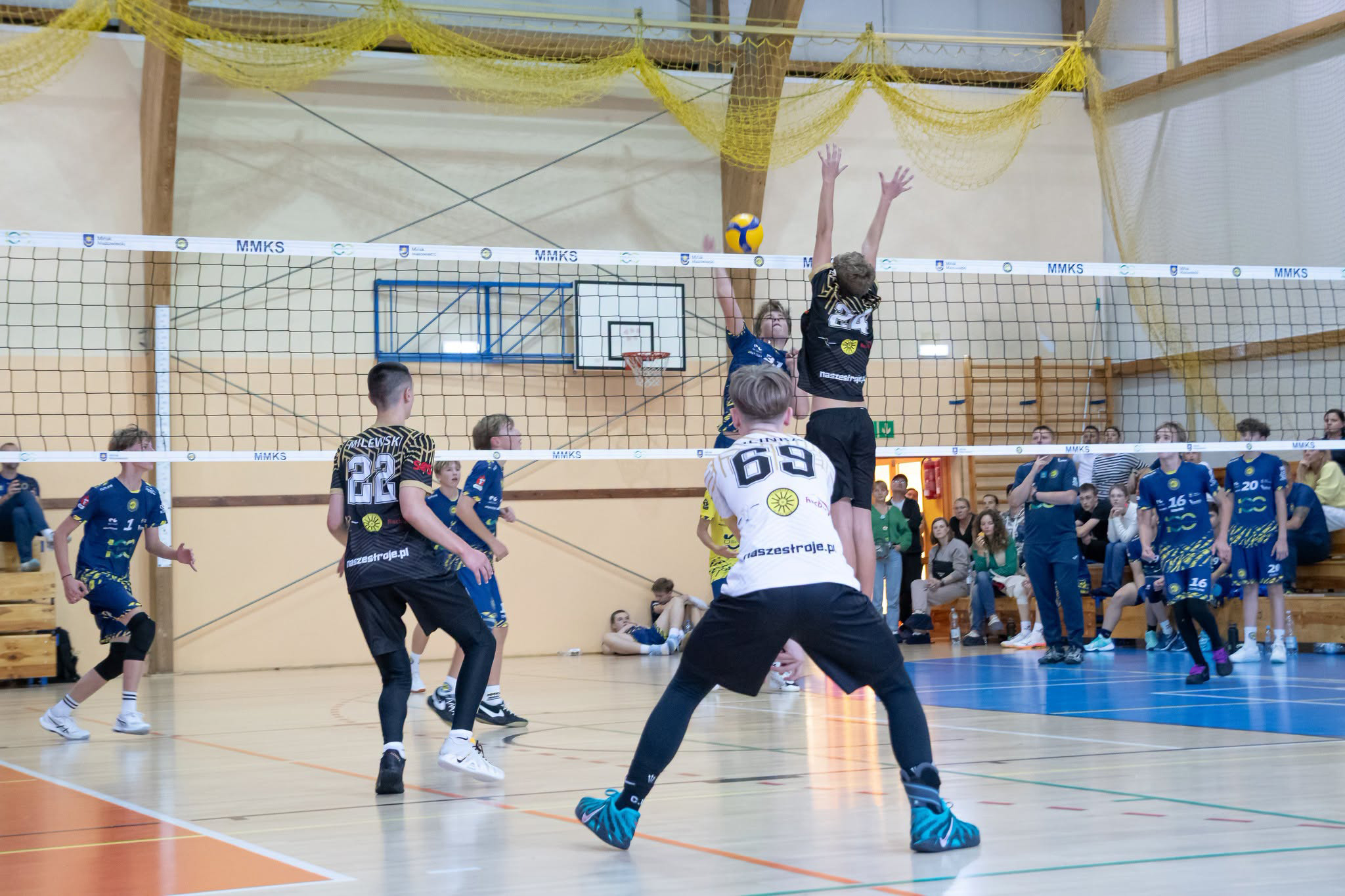 W hali sportowej dwóch siatkarzy skacze przy siatce, jeden z nich blokuje piłkę nad siatką. Wokół boiska są inni zawodnicy w czarnych i niebieskich strojach oraz widzowie na trybunach.