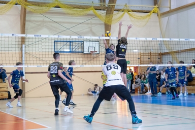 W hali sportowej dwóch siatkarzy skacze przy siatce, jeden z nich blokuje piłkę nad siatką. Wokół boiska są inni zawodnicy w czarnych i niebieskich strojach oraz widzowie na trybunach.