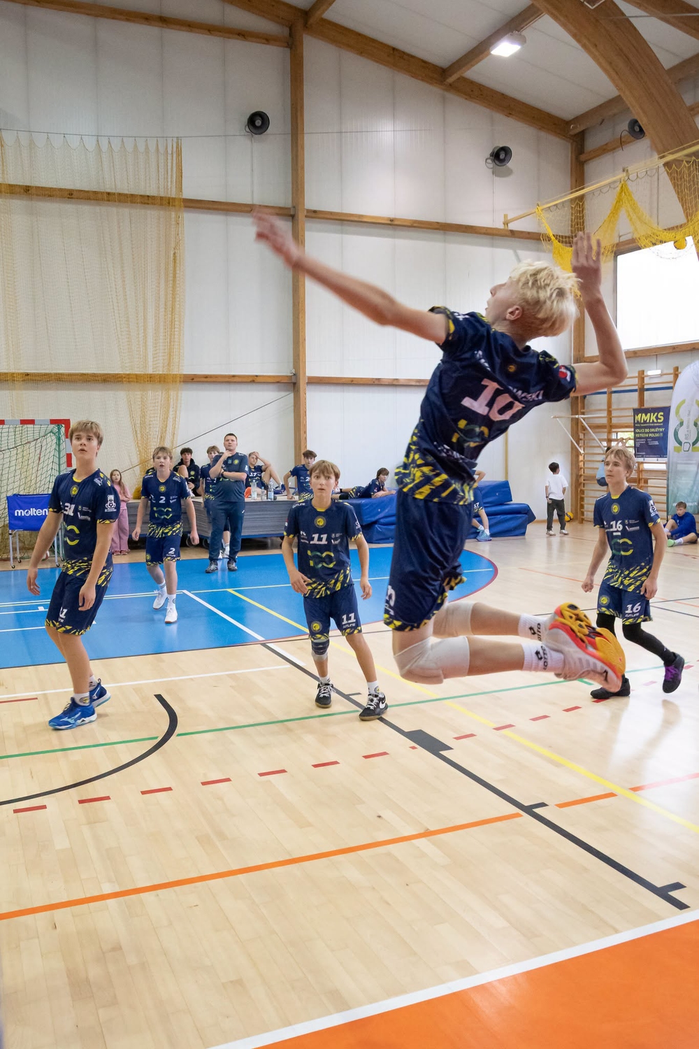 Chłopiec w ciemnym stroju skacze w powietrzu wykonując atak w hali sportowej. Kilku kolegów w podobnych strojach stoi na niebieskim boisku i obserwuje. W tle sędzia/trener przy stołem, drewniane belki konstrukcyjne i siatkarska siatka przy ścianie.