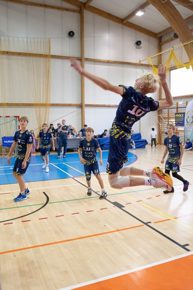 Chłopiec w ciemnym stroju skacze w powietrzu wykonując atak w hali sportowej. Kilku kolegów w podobnych strojach stoi na niebieskim boisku i obserwuje. W tle sędzia/trener przy stołem, drewniane belki konstrukcyjne i siatkarska siatka przy ścianie.