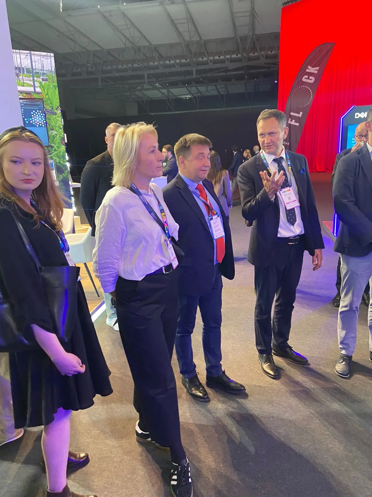 Grupa sześciu osób stojących wewnątrz hali konferencyjnej, trzy kobiety i trzech mężczyzn, większość ma identyfikatory na szyi, jedna osoba gestykuluje. W tle czerwony ekran i fragmenty stoisk targowych.