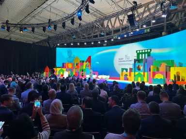 Wnętrze sali konferencyjnej z dużą liczbą siedzących osób zwróconych ku kolorowej scenie z prezentacją i kobietą przemawiającą. Na tle jasnoniebieskim wielokolorowe dekoracje sceniczne. Widoczne oświetlenie i elementy konstrukcyjne sufitu.