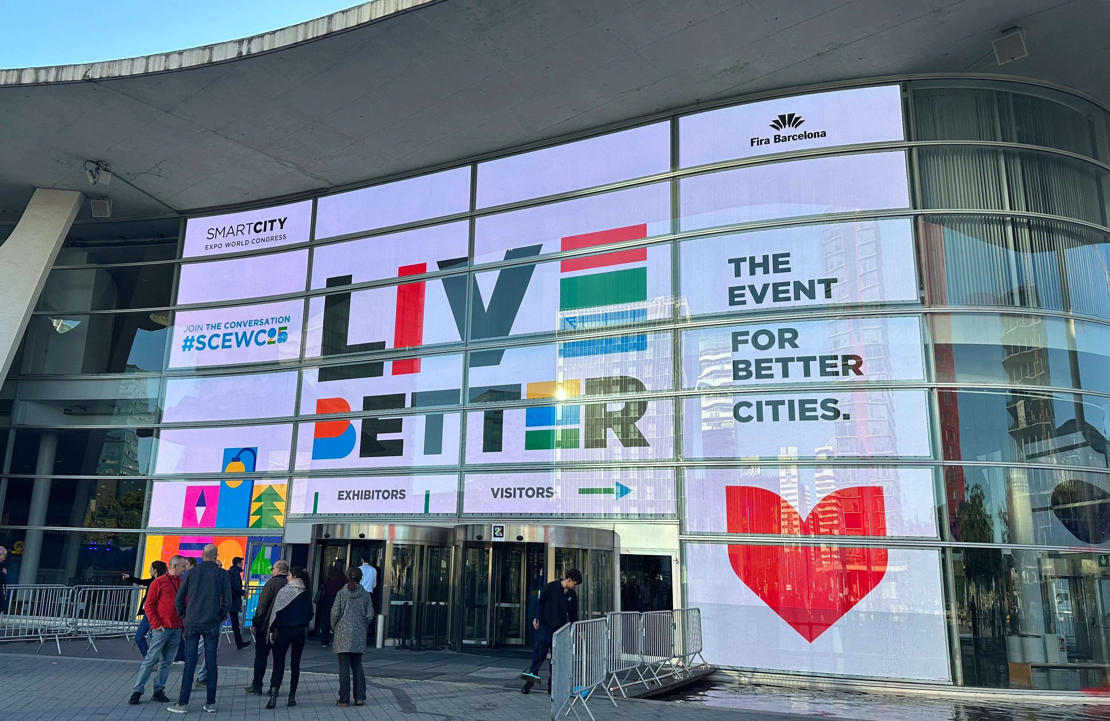 Grupa ludzi przed dużym budynkiem z przeszkloną fasadą. Na szklanej ścianie kolorowa grafika z napisem „LIVE BETTER” i tekstem „THE EVENT FOR BETTER CITIES.” oraz dużym czerwonym sercem. Widoczne tablice kierujące wystawców i odwiedzających. Słońce odbija się na szybach, jest dzień.