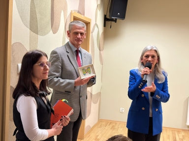 Trzy osoby stoją w jasnym pomieszczeniu z drewnianą podłogą. Po prawej kobieta w niebieskiej marynarce mówi do mikrofonu. Pośrodku mężczyzna w szarej marynarce trzyma małą ramkę z kolorowym motywem. Po lewej kobieta w okularach trzyma kilka notesów przy piersi, za nią jest ściana z dekoracyjnym wzorem.