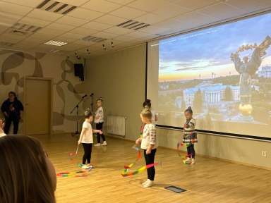 W sali z jasną drewnianą podłogą pięcioro dziewczynek tańczy z kolorowymi wstążkami przed dużym ekranem. Ekran pokazuje panoramę miasta i złotą rzeźbę. Po lewej stoi dorosła osoba przy drzwiach.