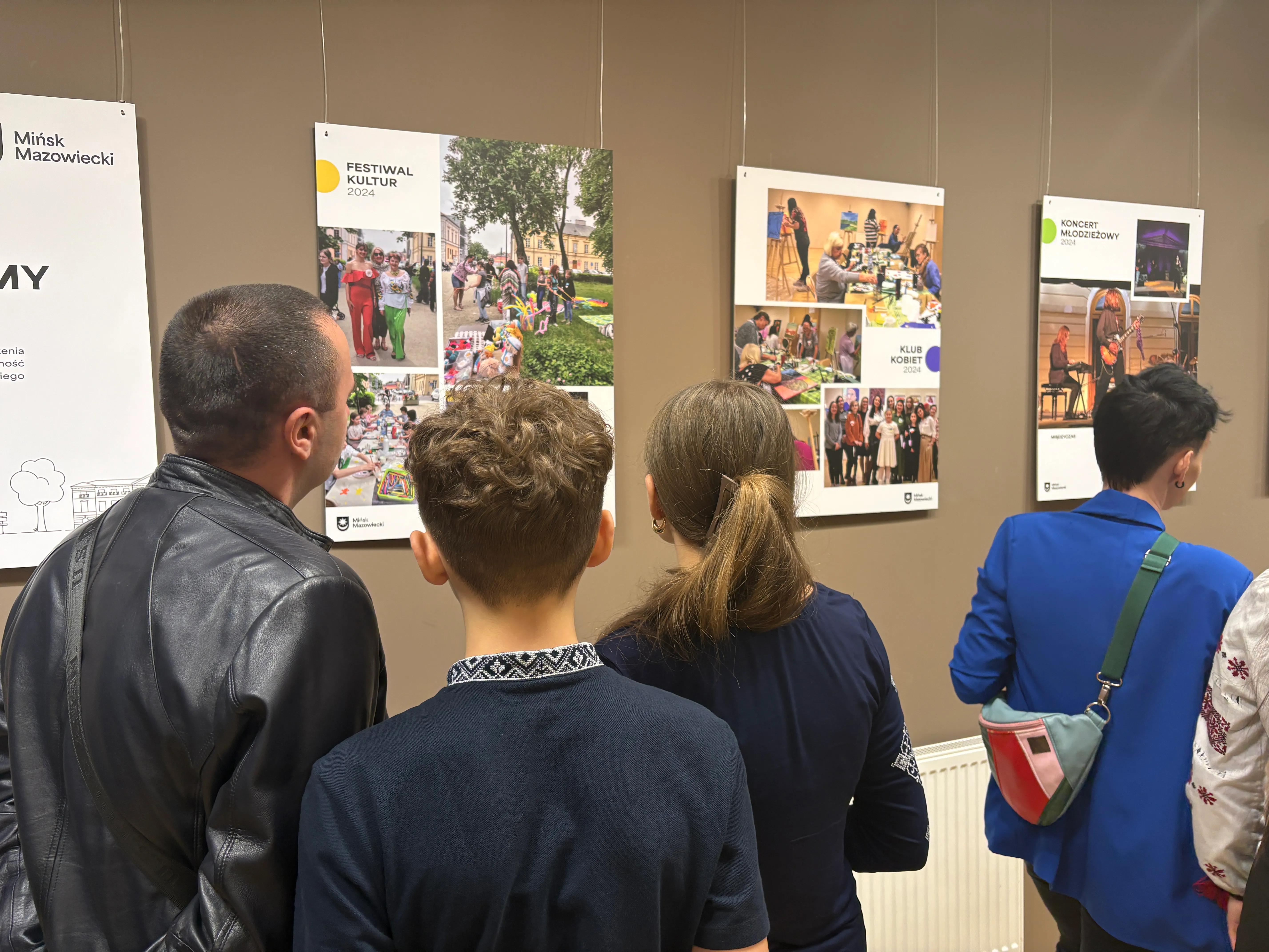 Pięć osób stoi plecami przed brązową ścianą z wiszącymi planszami wystawowymi. Sala jest wewnątrz. Lewa osoba ma czarną skórzaną kurtkę. Dziecko z krótkimi kręconymi włosami stoi obok. Kobieta z kucykiem stoi na środku. Po prawej osoba w niebieskiej marynarce ma kolorową torebkę przewieszoną przez ramię. Pod planszami widoczny jest biały grzejnik.