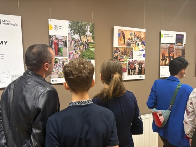 Pięć osób stoi plecami przed brązową ścianą z wiszącymi planszami wystawowymi. Sala jest wewnątrz. Lewa osoba ma czarną skórzaną kurtkę. Dziecko z krótkimi kręconymi włosami stoi obok. Kobieta z kucykiem stoi na środku. Po prawej osoba w niebieskiej marynarce ma kolorową torebkę przewieszoną przez ramię. Pod planszami widoczny jest biały grzejnik.