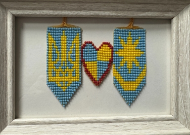 Trzy zawieszone ozdoby z małych koralików na białym tle w jasnej drewnianej ramie: po bokach dwie niebiesko‑żółte chorągiewki z zakończeniem w szpic, pośrodku serce wypełnione na przemian niebieskimi i żółtymi koralikami obramowane czerwonymi.