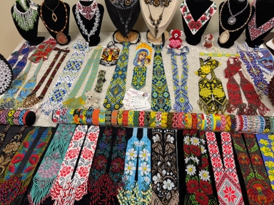 Stół wewnątrz z wieloma ręcznie robionymi koralikowymi naszyjnikami, krawatkami i bransoletkami ułożonymi w rzędach. W tylnej części stojaki z czarnymi i beżowymi manekinami prezentują większe naszyjniki. Wyraźne, kolorowe wzory geometryczne i kwiatowe na jasnym tle materiału.