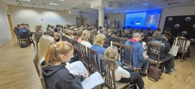 Wnętrze sali konferencyjnej z podświetlonym ekranem z frontu. W kilkunastu rzędach drewnianych krzeseł siedzi około pięćdziesięciu młodych osób. Na ekranie widoczne symbole Unii Europejskiej i Ukrainy oraz wykład prowadzony przez kobietę. Ściany beżowe, podłoga jasne drewno, sufit z białych paneli.