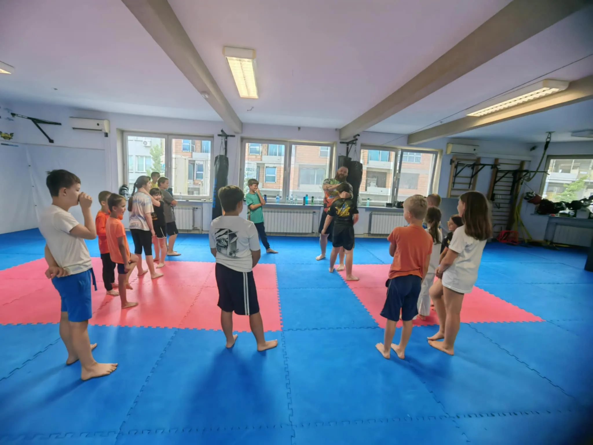 Grupa dzieci w wieku szkolnym stoi na matach do sportów walki podczas zajęć prowadzonych przez instruktora w sali treningowej.