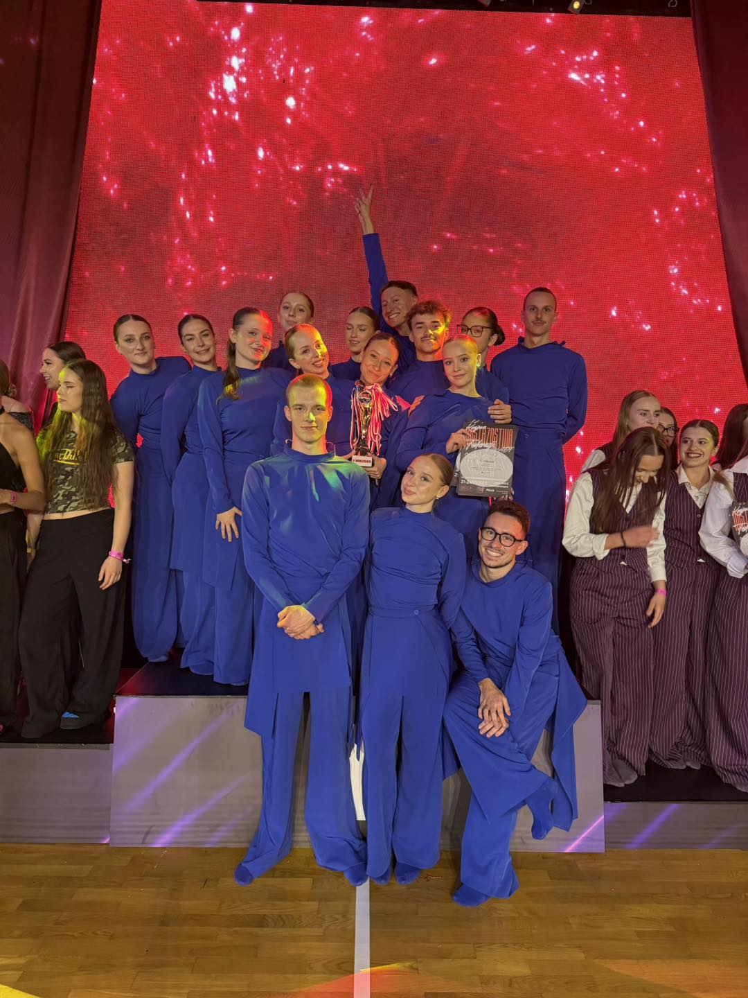 Grupa kilkunastu osób stoi na scenie. Wszyscy mają długie, jednolite niebieskie stroje. Jedna osoba trzyma puchar, inna trzyma dyplom. Za grupą świeci duże czerwone, migające tło LED. Wnętrze sali i drewniana podłoga są widoczne.