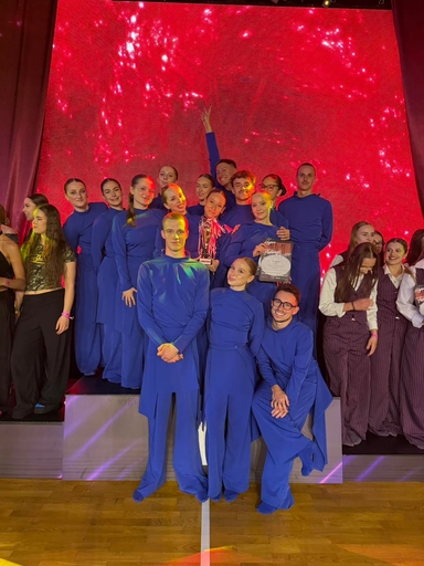 Grupa kilkunastu osób stoi na scenie. Wszyscy mają długie, jednolite niebieskie stroje. Jedna osoba trzyma puchar, inna trzyma dyplom. Za grupą świeci duże czerwone, migające tło LED. Wnętrze sali i drewniana podłoga są widoczne.