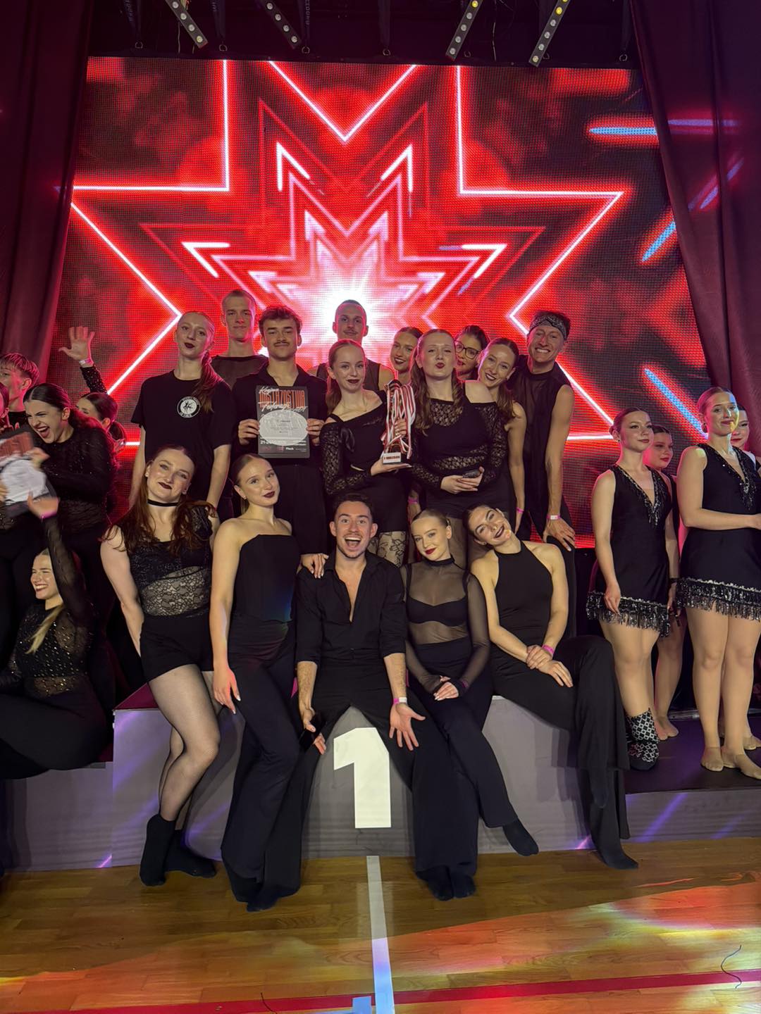 Grupa kilkunastu tancerzy stoi na scenie w czarnych strojach. Centralnie trzy osoby siedzą na podium z oznaczeniem "1", jedna trzyma puchar, inna dyplom. W tle duża, jasnoczerwona neonowa gwiazda na ekranie LED.