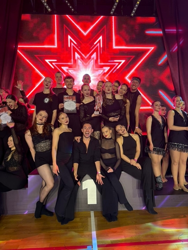 Grupa kilkunastu tancerzy stoi na scenie w czarnych strojach. Centralnie trzy osoby siedzą na podium z oznaczeniem "1", jedna trzyma puchar, inna dyplom. W tle duża, jasnoczerwona neonowa gwiazda na ekranie LED.