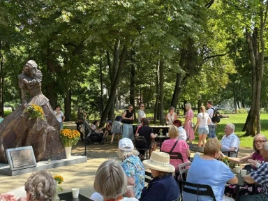 Brązowa rzeźba kobiety w historycznym ubraniu obok grupy ludzi siedzących i stojących w parku pod drzewami.