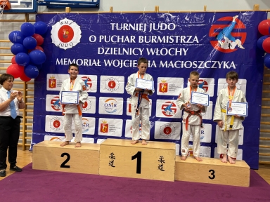 Czterech chłopców w judogach stoi na podium z dyplomami i medalami w hali sportowej podczas turnieju judo.
