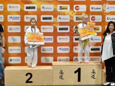 Dwie dziewczynki w białych judogach stoją na podium na miejscach pierwszym i drugim. Dziewczynka na pierwszym miejscu trzyma puchar i dyplom, ma żółty pas. Dziewczynka na drugim miejscu trzyma dyplom, ma biały pas. W tle pomarańczowa ścianka z logo sponsorów. Po prawej stoi kobieta w białej koszuli i czarnej kamizelce. Po lewej częściowo widoczna osoba dorosła.