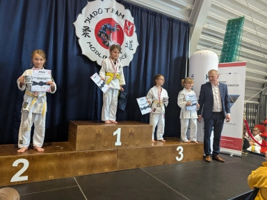 Cztery dziewczynki w białych judogach stoją na podium z trzema stopniami, trzymając dyplomy i medale. Po prawej stoi mężczyzna w garniturze. W tle duży baner z napisem MUDO TEAM MODLICE.