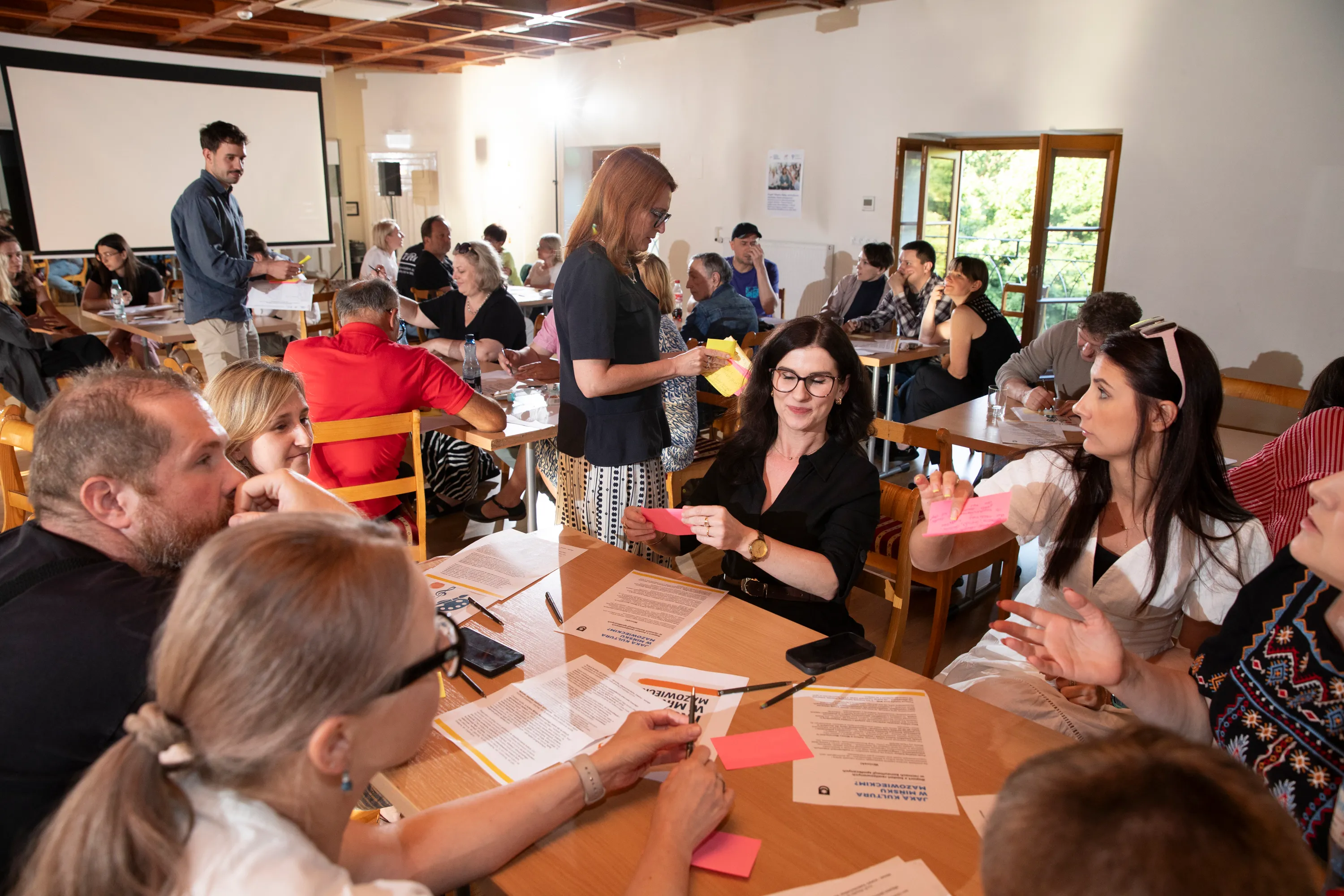 Wnętrze sali konferencyjnej z kilkoma grupami ludzi siedzącymi przy stołach podczas warsztatów, kobieta w czarnej bluzce trzyma kartkę, mężczyzna z brodą i inne osoby rozmawiają, jest dzień, za oknem zieleń.