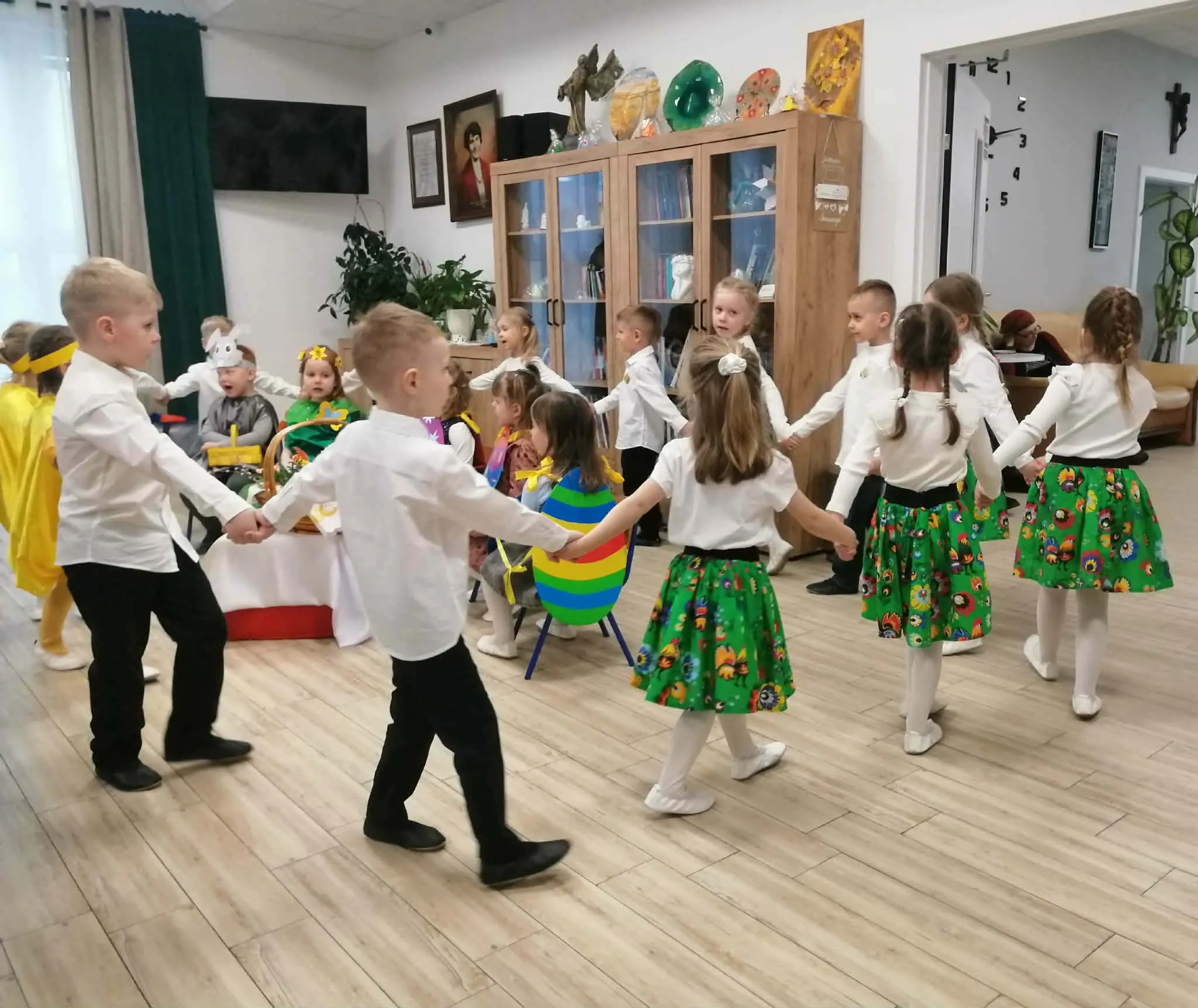Kilku dzieci trzyma się za ręce i tańczy w kole na jasnej drewnianej podłodze. Dziewczynki mają białe bluzki i zielone spódniczki w kolorowy wzór, chłopcy białe koszule i czarne spodnie. W tle szafka z półkami, dekoracje i okno z zasłonami, wnętrze oświetlone światłem dziennym.