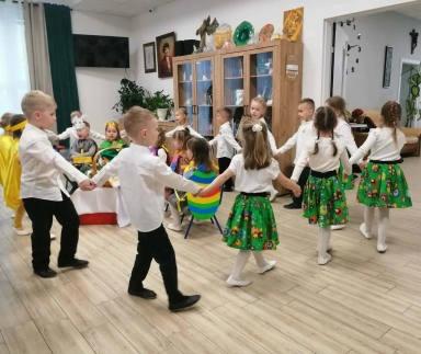 Kilku dzieci trzyma się za ręce i tańczy w kole na jasnej drewnianej podłodze. Dziewczynki mają białe bluzki i zielone spódniczki w kolorowy wzór, chłopcy białe koszule i czarne spodnie. W tle szafka z półkami, dekoracje i okno z zasłonami, wnętrze oświetlone światłem dziennym.