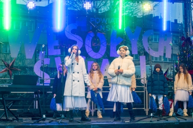 Sześć dzieci stoi na scenie podczas występu na zewnątrz, dwie dziewczynki na przodzie trzymają mikrofony, pozostałe dzieci mają niebieskie pompony. Dzieci są ubrane w jasne ubrania i kurtki zimowe. Za nimi znajduje się duży napis „WESOŁYCH ŚWIĄT” i kolorowe oświetlenie. W tle widoczne są budynki i choinka udekorowana światełkami.