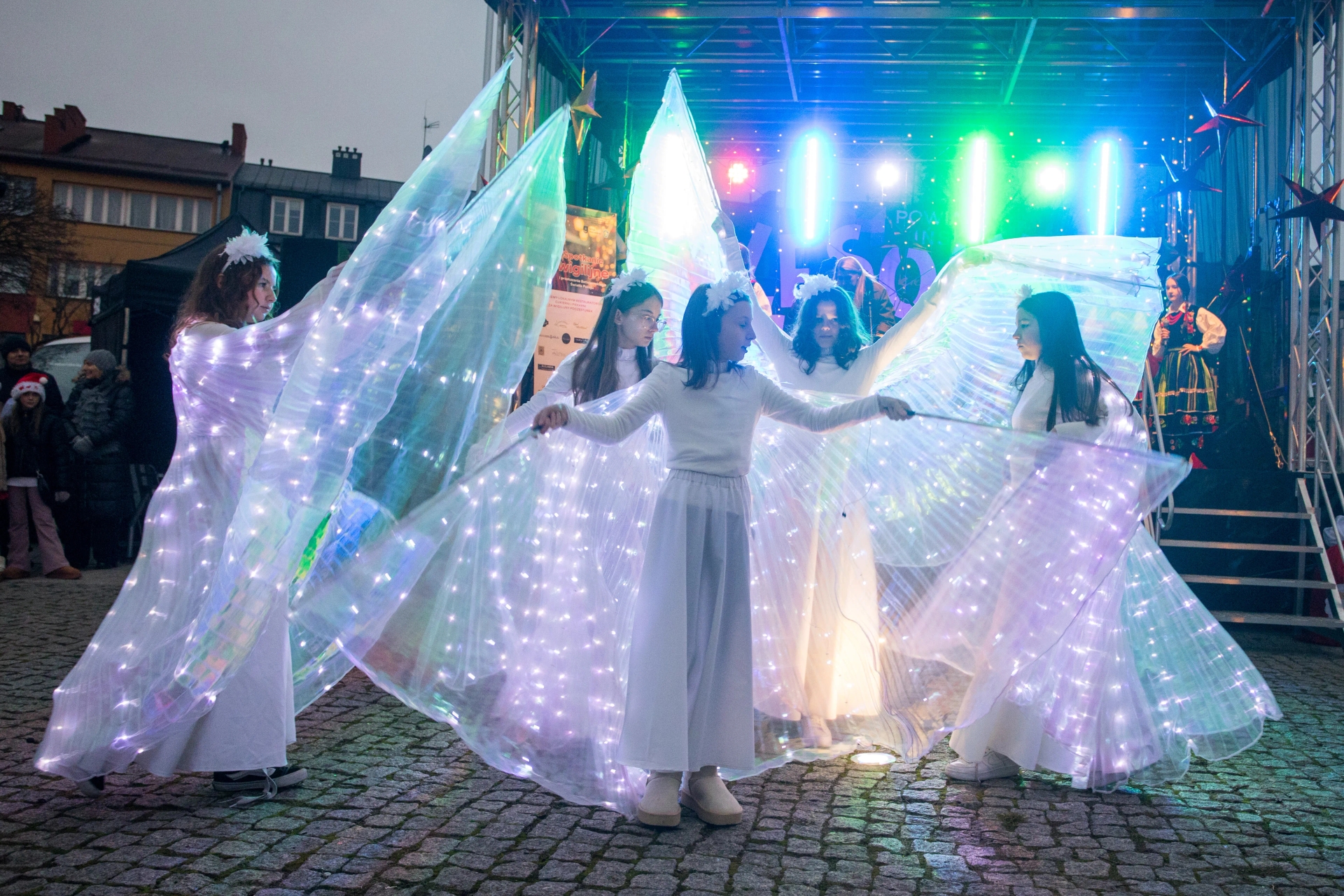 Pięć dziewcząt w białych strojach z dużymi, podświetlanymi skrzydłami, tańczących na otwartym placu w pobliżu sceny z kolorowymi światłami, wieczór.