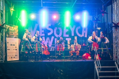 Na scenie stoi i siedzi siedmioro osób w tradycyjnych, kolorowych strojach ludowych. Pięć osób śpiewa, jedna gra na skrzypcach, jedna na akordeonie, a jedna na wiolonczeli. W tle duży napis „WESOŁYCH ŚWIĄT” oraz kolorowe światła. Po prawej stronie jest schodek na scenę, po lewej baner z napisem i logo. Scena jest udekorowana ciemnym tłem i wiszącymi gwiazdami.