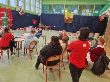 Grupa osób siedzi na krzesłach w sali gimnastycznej, jedna osoba stoi twarzą do grupy, na ścianie dekoracje z balonów i pakunki, w tle kosze do koszykówki oraz zasłony z ciemnego materiału.