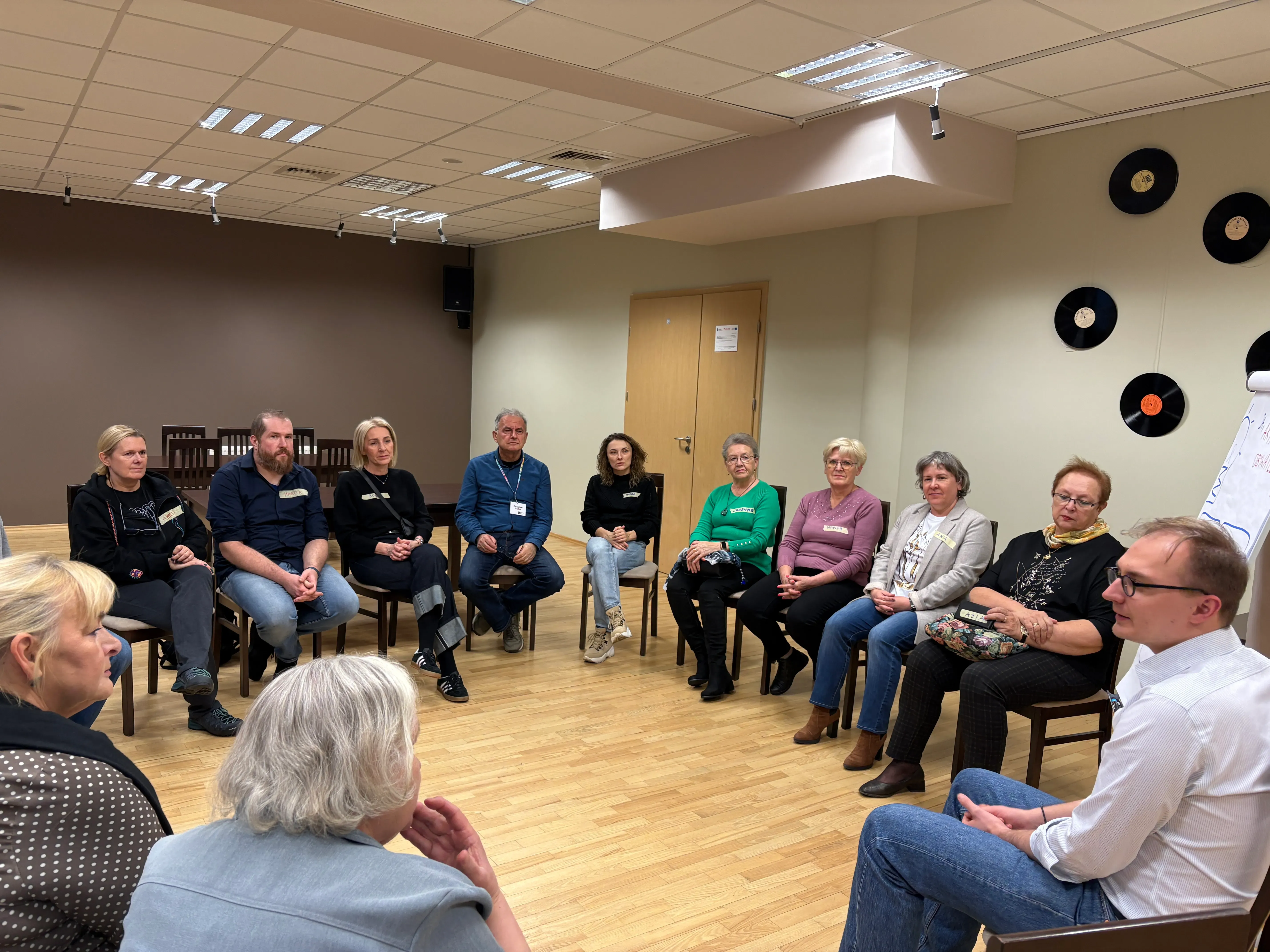 Grupa dwunastu osób siedzi na krzesłach w kole w jasnym pomieszczeniu z drewnianą podłogą. Na ścianie po prawej stronie wiszą czarne płyty winylowe. W tle znajdują się drewniane drzwi.