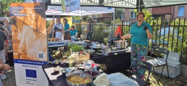 Kobieta w kolorowych spodniach i turkusowej koszulce stoi za stołem z naczyniami i składnikami do gotowania na zewnątrz w ciągu dnia pod namiotem. Obok stoi baner z informacjami o UE, a w tle kilka osób rozmawia i robi zdjęcia.