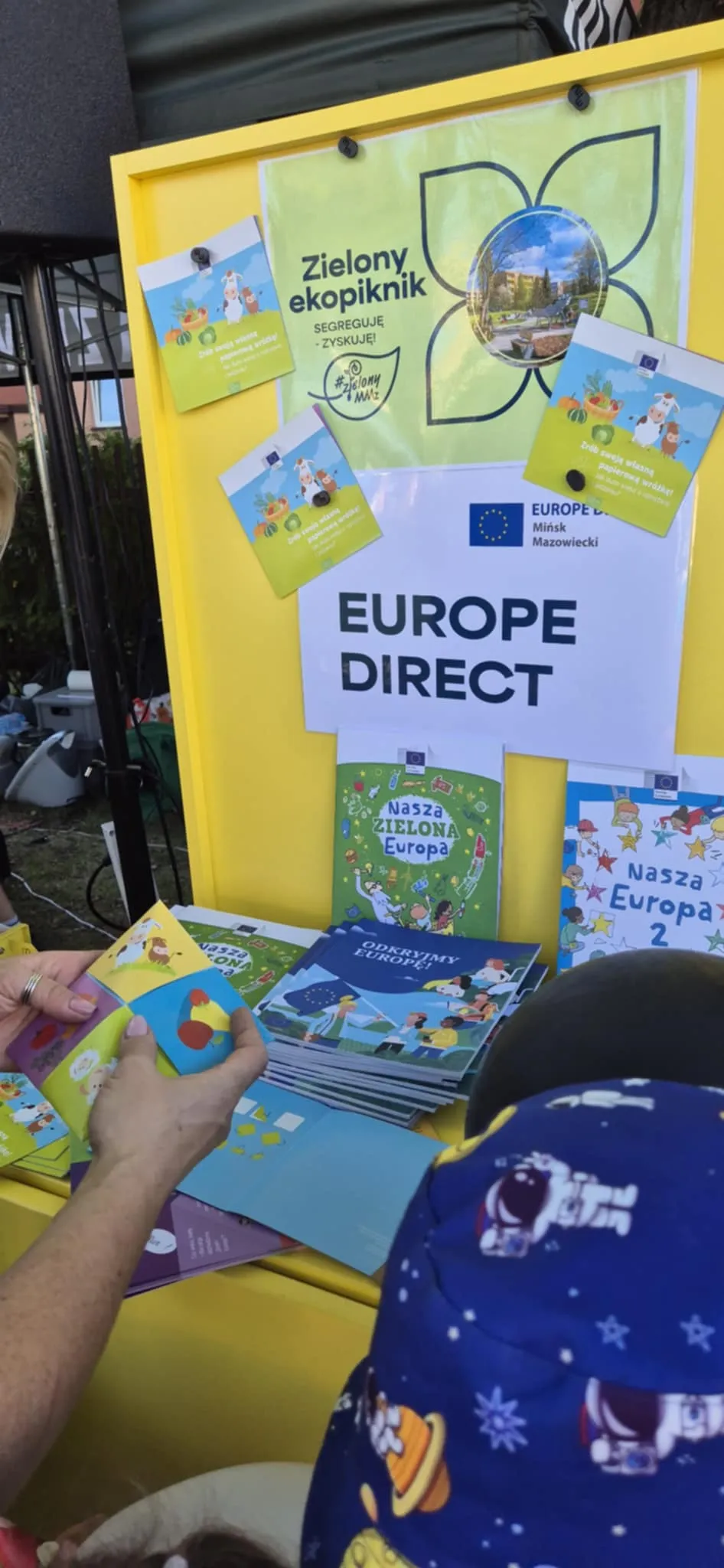 Stoisko z żółtą ścianką, na której wiszą ulotki i plakaty związane z ekologią i Unią Europejską. Na ścianie duży napis "EUROPE DIRECT". Ręce osoby oglądającej broszurę, obok dziecko w niebieskiej czapce ozdobionej wzorami astronautów i planet.